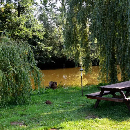 Ein Holztisch und eine Bank stehen auf einem Grasfeld in der Nähe eines Flusses mit gelblichem Wasser. Ein Lampenpfahl befindet sich auf dem Grasfeld in der Nähe des Flusses. Bäume stehen auf beiden Seiten des Flusses.