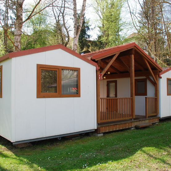 Eine weiße Hütte mit einem roten Dach befindet sich in einem Grasbereich, mit mehreren Bäumen im Hintergrund. Die Hütte verfügt über eine Holzveranda mit Geländern, ein Fenster mit Jalousien und ein Feuerlöscherschild.