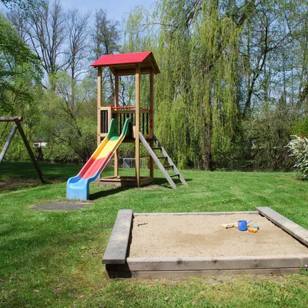 Ein Spielplatz mit einem bunten Rutschen, einem Sandkasten und einer Schaukel in einem von Bäumen umgebenen Rasenbereich.