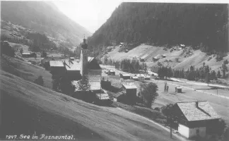 Ein Schwarz-Weiß-Foto von 1947 zeigt ein kleines Dorf in Paznaun, Österreich, mit einer Kirche und mehreren Häusern in einer Berglandschaft.
