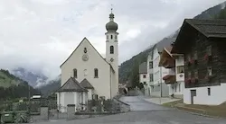 Eine kleine Kirche mit einem Turm und einer Uhr steht in einem Dorf mit Häusern und Bergen im Hintergrund.