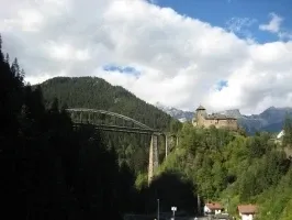 Eine Brücke über einen Fluss mit einer Burg auf einem Hügel und Bergen im Hintergrund.