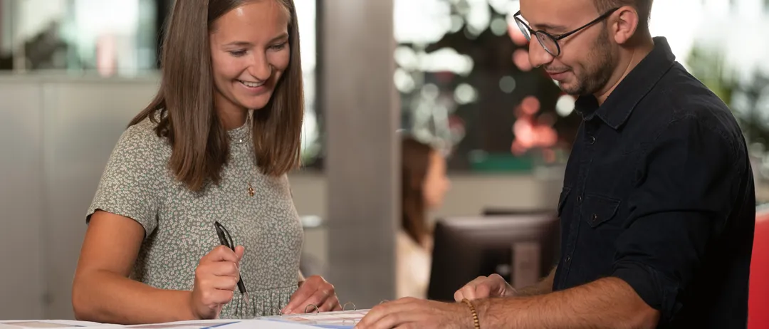 Eine Frau und ein Mann stehen an einem Schreibtisch, lächeln und schauen sich Papiere an. Die Frau hält einen Stift und scheint ein Dokument zu unterschreiben.