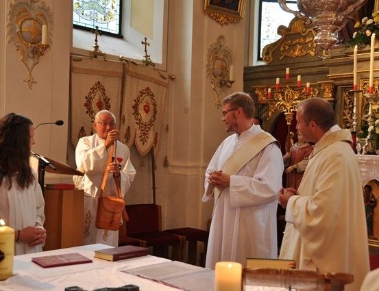 Eine Gruppe von Priestern und einer Frau versammelt sich in einer Kirche, wobei ein Priester eine Tasche und ein anderer ein Mikrofon hält. Sie stehen vor einem Tisch mit Kerzen und Büchern.