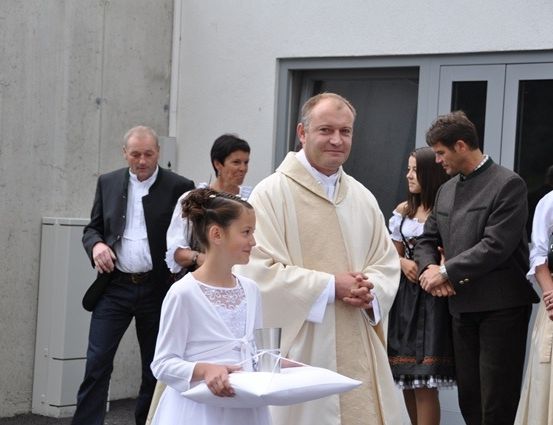 Ein Priester steht neben einem jungen Mädchen, das ein Kissen hält, mit mehreren Personen in traditioneller Kleidung im Hintergrund.
