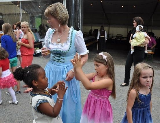 Eine Frau in traditioneller bayerischer Tracht interagiert mit jungen Mädchen bei einer Veranstaltung im Freien, während andere Menschen im Hintergrund herumlaufen.