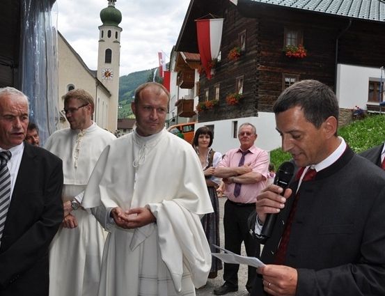 Ein Mann in einem schwarzen Anzug hält ein Mikrofon und spricht zu einer Gruppe von Menschen, darunter zwei Priester in weißen Gewändern, die vor einem Gebäude mit Turm und Fahnen stehen.