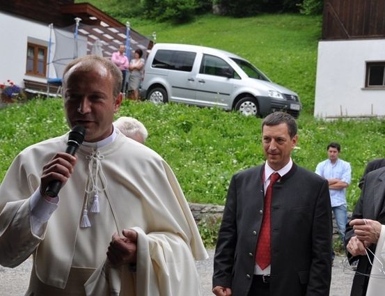 Ein Priester in einer weißen Robe spricht in ein Mikrofon, während ein Mann in einem schwarzen Anzug und roter Krawatte in der Nähe steht und andere im Hintergrund beobachten.