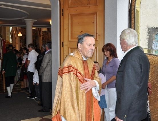 Ein Priester in einer goldenen Robe steht in einer Kirche und begrüßt einen Mann im Anzug. Dahinter wartet eine Gruppe von Menschen in der Schlange.
