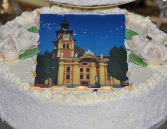 Ein weißer Kuchen mit einem Foto einer Kirche darauf, verziert mit weißen Rosen und grünen Blättern.