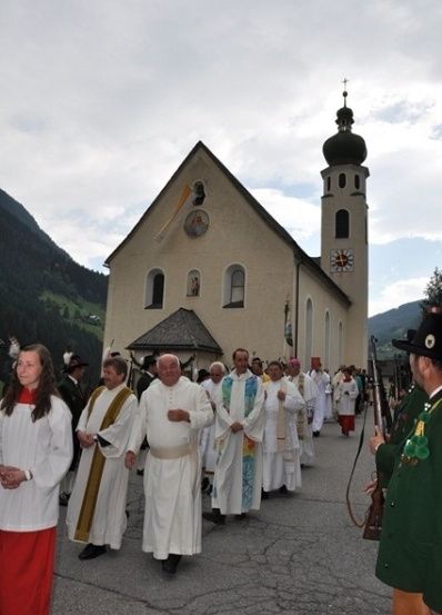 Eine Prozession von Priestern in weißen Gewändern geht vor einer Kirche mit Turm und Uhr.