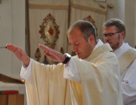 Ein Priester in einer weißen Robe betet mit erhobenen Händen in einer Kirche. Ein anderer Priester steht hinter ihm.