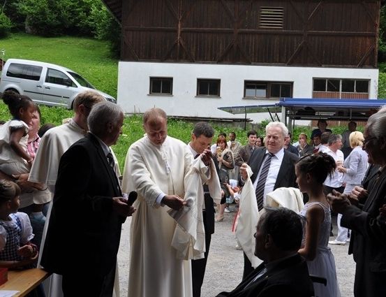 Ein Priester in einer weißen Robe führt eine Zeremonie vor einer Menge durch, mit einem Gebäude und einem Van im Hintergrund.