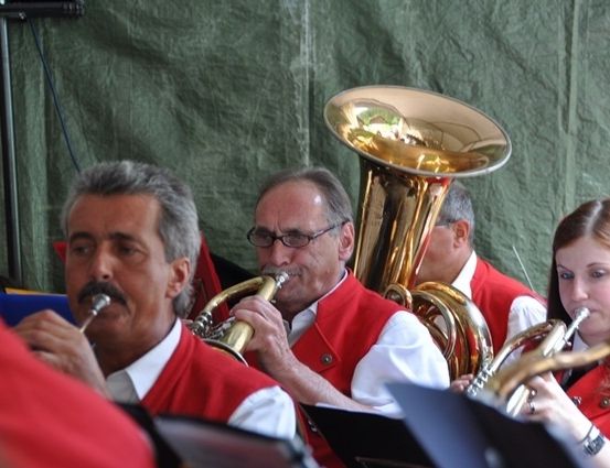 Eine Gruppe von Musikern in roten Westen spielt Blechblasinstrumente, darunter Tubas und Trompeten, in einer Außenumgebung.