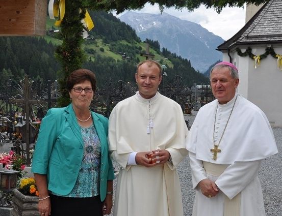Drei Personen, darunter zwei Priester und eine Frau, stehen vor einer Kirche mit Bergkulisse.