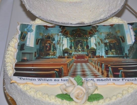 Ein Kuchen mit einem Foto des Kircheninneren, mit weißer Zuckergussverzierung, einer weißen Rose und einem deutschen Satz am Rand.