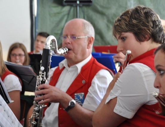 Ein Mann und eine Frau in roten Westen spielen Klarinetten in einer Band, mit anderen Bandmitgliedern und Notenständern im Hintergrund.