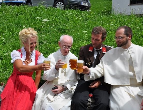 Eine Gruppe von Menschen, darunter zwei Priester, sitzen auf einem Rasen und halten Gläser mit Bier. Eine Frau in einem roten Kleid lächelt.