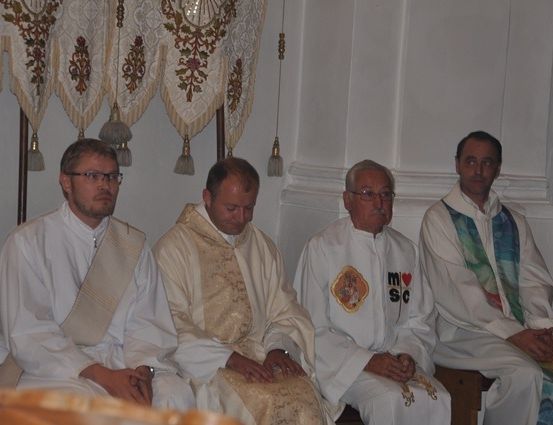 Vier Männer in weißen Gewändern sitzen in einer Reihe, möglicherweise Priester, einer mit Brille und einer mit einem bunten Schal. Sie sind in einem Raum mit dekorativen Vorhängen.