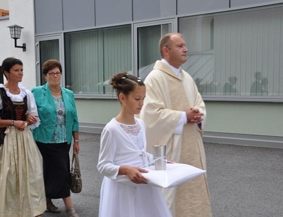 Ein junges Mädchen in einem weißen Kleid hält ein Kissen, während ein Priester neben ihr steht, und zwei Frauen in traditioneller Kleidung stehen hinter ihnen.