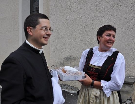 Ein lächelnder Priester in einer schwarzen Robe hält eine Schachtel mit Gebäck in der Hand, während er neben einer Frau in traditioneller bayerischer Kleidung steht.