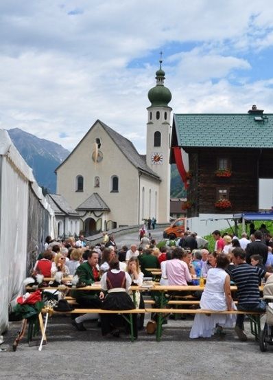 Eine Gruppe von Menschen sitzt an Tischen vor einer Kirche mit einer grünen Kuppel und einem Glockenturm.