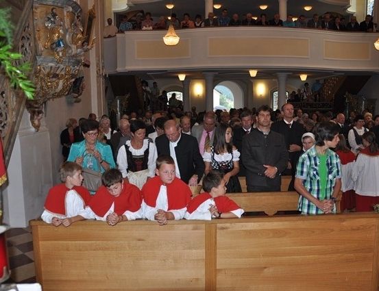 Eine Gruppe von Kindern in roten und weißen Gewändern sitzt auf einer Holzbank in einer Kirche, mit Erwachsenen hinter ihnen.