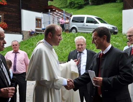 Ein Priester in einer weißen Robe schüttelt einem Mann in einem schwarzen Anzug die Hand, während andere hinter ihnen auf einer Rasenfläche stehen.