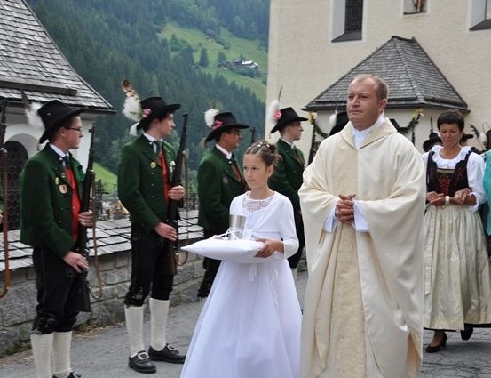 Ein Priester und ein junges Mädchen in einem weißen Kleid stehen vor einer Kirche mit traditionellen Wächtern in grünen Uniformen.