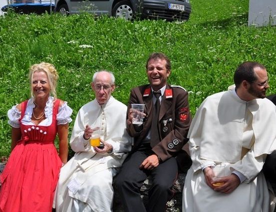 Eine Gruppe von Menschen, darunter zwei Priester, sitzen auf einer Steinmauer, lächeln und halten Getränke in der Hand. Ein Auto ist hinter ihnen geparkt.