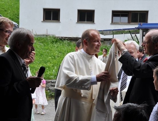 Ein Priester wird von zwei Männern bei der Entfernung seiner Veste unterstützt, während ein Mann ein Mikrofon hält und ein junges Mädchen in der Nähe steht.