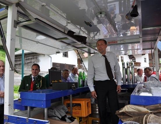 Ein Mann in Anzug steht in einer mobilen Bar, umgeben von Leuten und Flaschen auf der Theke.