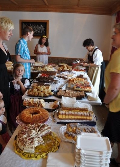 Eine Gruppe von Menschen versammelt sich um einen langen Tisch, der mit verschiedenen Desserts wie Kuchen, Gebäck und Brot gefüllt ist. Einige stehen, andere sitzen. Eine Frau in einem gelben Shirt lächelt.