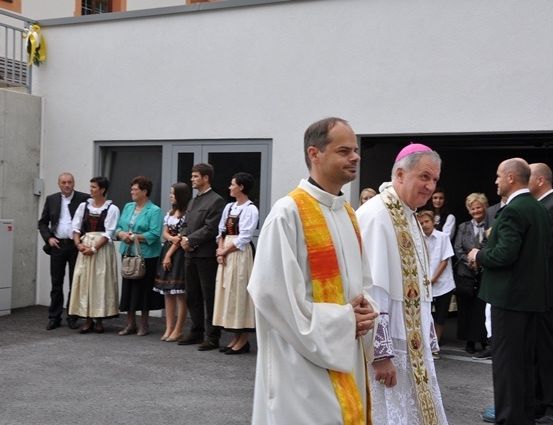 Zwei Priester stehen vor einer Gruppe von Menschen, möglicherweise während einer religiösen Zeremonie. Die Priester tragen traditionelle religiöse Kleidung, und die Menschen hinter ihnen sind in formeller Kleidung gekleidet.