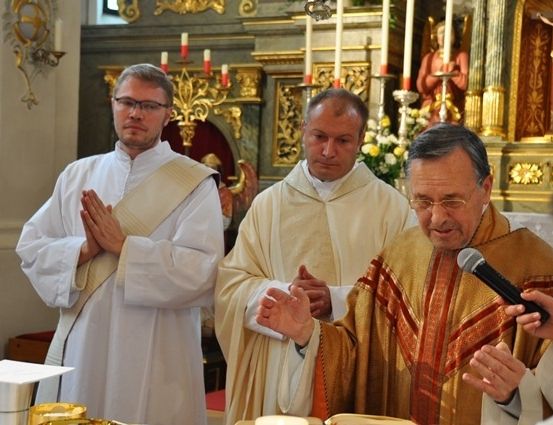 Drei Priester stehen vor einem Altar, einer hält ein Mikrofon, wahrscheinlich während einer religiösen Zeremonie. Der Priester in der Mitte trägt eine Brille und eine Stola. Hinter ihnen ist ein goldener Altar mit Kerzen und einer Statue zu sehen.