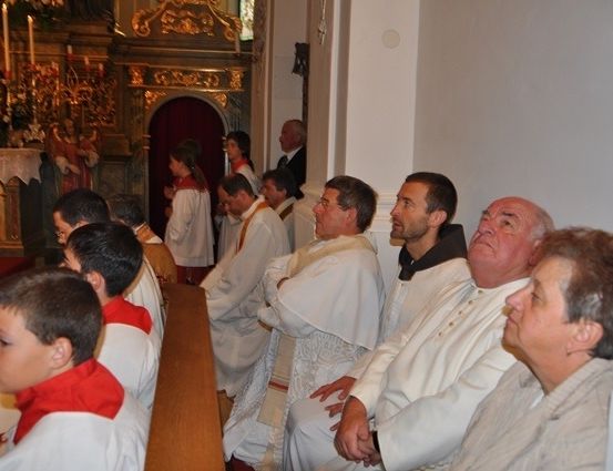 Eine Gruppe von Menschen in weißen Gewändern sitzt in einer Kirche. Einige tragen Brillen. Hinter ihnen steht ein Altar mit Kerzen und einer Statue.