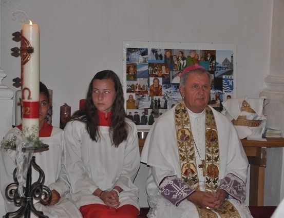 Ein Priester und zwei junge Menschen in weißen Gewändern sitzen vor einer Kerze und einer Collage von Fotos an einer Wand.