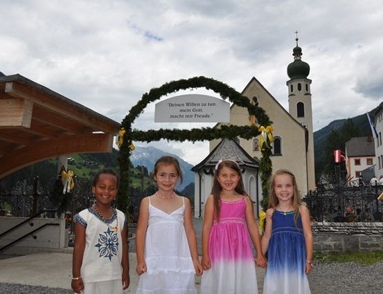 Vier junge Mädchen stehen vor einer Kirche mit einem dekorativen Bogen und einem Schild, das 'Deinen Willen zu tun, mein Gott, macht mir Freude.' hinter ihnen sind Berge und Gebäude zu sehen.
