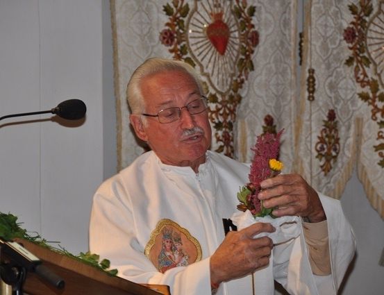 Ein älterer Priester in einer weißen Robe hält einen Blumenstrauß, während er an einer Kanzel steht.