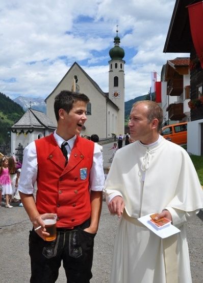 Ein Priester und ein Mann in traditioneller Kleidung stehen vor einer Kirche. Der Mann hält ein Bier und eine Zigarette.
