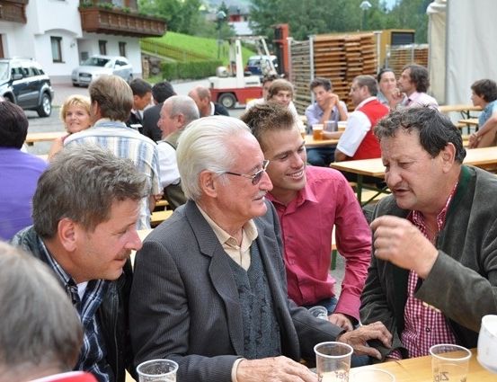Eine Gruppe von Menschen sitzt an einem Tisch in einem Restaurant im Freien. Sie führen ein Gespräch und scheinen sich zu amüsieren. Auf dem Tisch stehen Tassen und Teller. Im Hintergrund sitzen andere Leute an Tischen, und einige stehen. In der Ferne sind auch Autos geparkt.