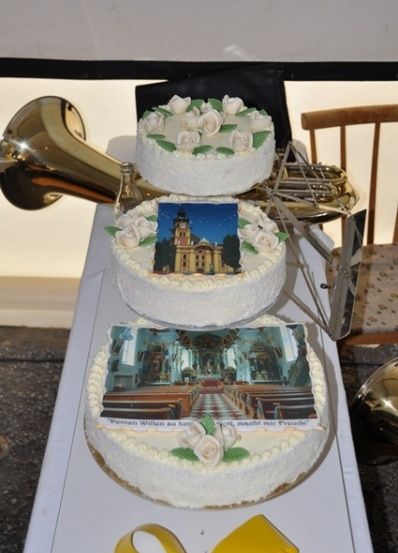 Drei weiß glasierte Kuchen mit Bildern einer Kirche darauf stehen auf einem Tisch mit einem Musikhorn und einem Stuhl im Hintergrund.