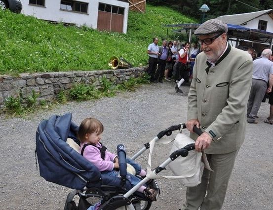 Ein älterer Mann schiebt einen Kinderwagen mit einem kleinen Mädchen darin. Dahinter steht eine Gruppe von Menschen in der Nähe eines Gebäudes mit einer Tuba.