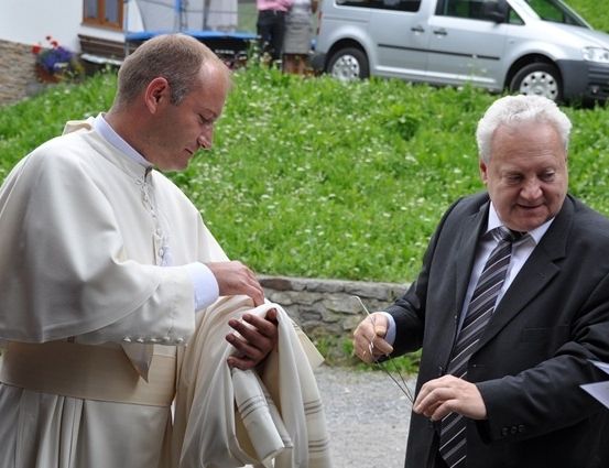 Ein Priester legt einem Mann in einem Anzug eine Stola um den Hals, mit einem Auto und einem Gebäude im Hintergrund.