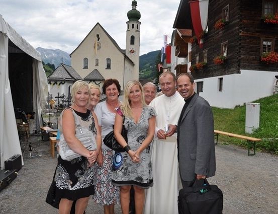 Eine Gruppe von Menschen, einschließlich eines Priesters, steht vor einer Kirche mit einer grünen Kuppel und lächelt für ein Foto. Hinter ihnen sind ein weißes Zelt und eine Holzbank zu sehen.