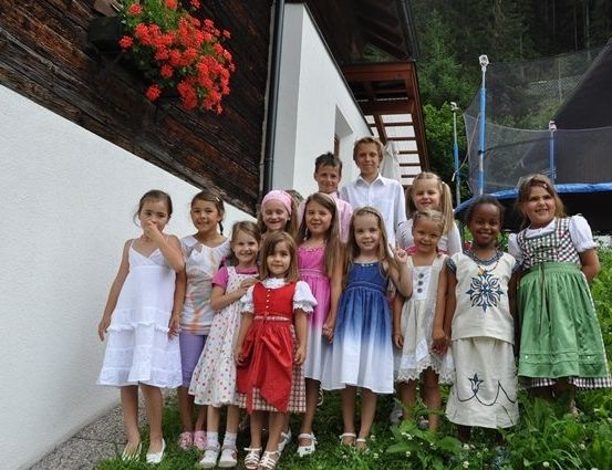 Eine Gruppe von Kindern und zwei Erwachsenen steht vor einem Haus mit einem roten Blumenkasten an der Wand. Die Kinder tragen verschiedene Kleider und Schuhe.