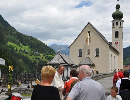 Eine Gruppe von Menschen versammelt sich vor einer Kirche mit einer grünen Kuppel und einem Glockenturm. Die Kirche ist von einem Friedhof mit Kreuzen und einem Kreuz auf dem Hügel umgeben.