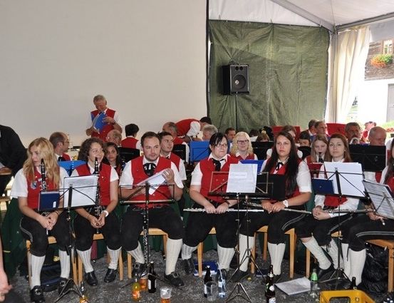Eine Gruppe von Musikern in roten Westen und schwarzen Hosen sitzen in Stühlen und spielen ihre Instrumente in einem Zelt.