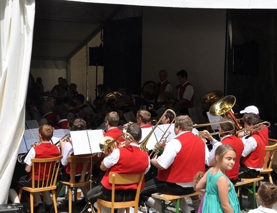Ein Blasorchester spielt auf der Bühne unter einem Zelt, mit Musikern, die Posaunen und Tuben spielen. Zuschauer, darunter Kinder, sitzen in Stühlen vor der Bühne.
