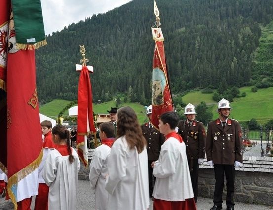 Eine Gruppe von Menschen in weißen Roben und roten Umhängen steht vor einer Steinmauer mit Fahnen und Bergen im Hintergrund.